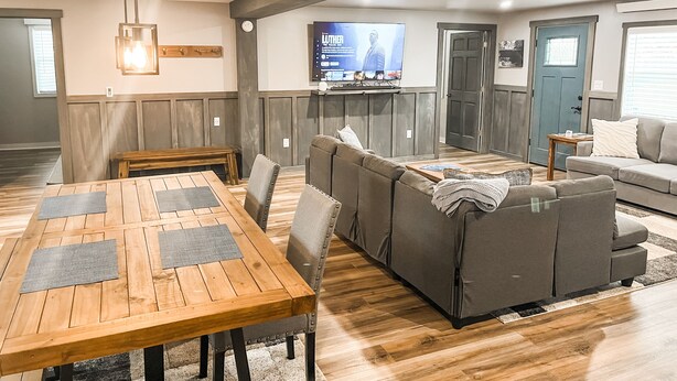Un salon moderne avec une table à manger en bois, un canapé gris et un téléviseur fixé au mur.