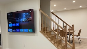 A modern living room with a flat-screen TV mounted on the wall, a wooden staircase, and a dining area with chairs. - One of the kind fully loaded house located walking distance from cozy lake (Albrightsville)
