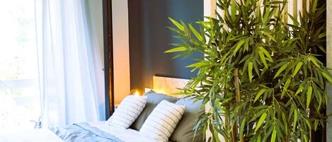 A bedroom with a bed, pillows, a bedside table, a lamp, and a large bamboo plant.