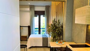 Ein kleines Hotelzimmer mit Bett, Esstisch und Stühlen, einer Küchenzeile mit Spüle und einem großen Fenster mit Vorhängen.