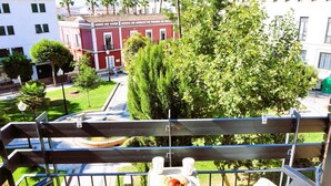 Ein Balkon mit Sitzgelegenheit im Freien, einem Tisch mit Frühstücksgebäck und Blick auf ein rotes Gebäude und Bäume.