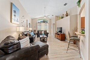Un salon spacieux avec un canapé, une table basse et un téléviseur. Il y a également un coin repas avec une chaise et une table. La pièce est dotée d’un plancher en bois et d’un ventilateur de plafond.