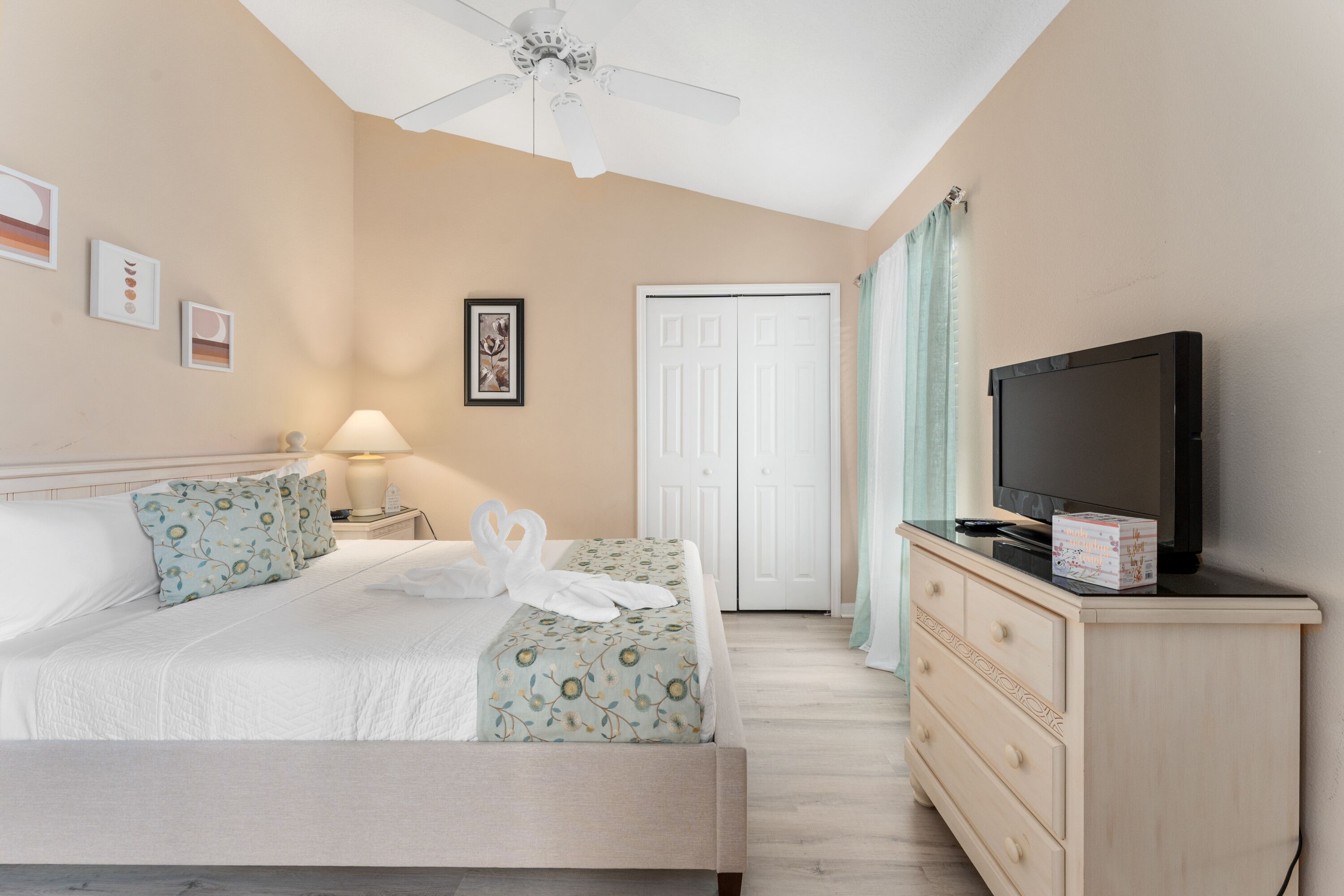 A hotel room with a bed, a TV, a dresser, a ceiling fan, and a door.