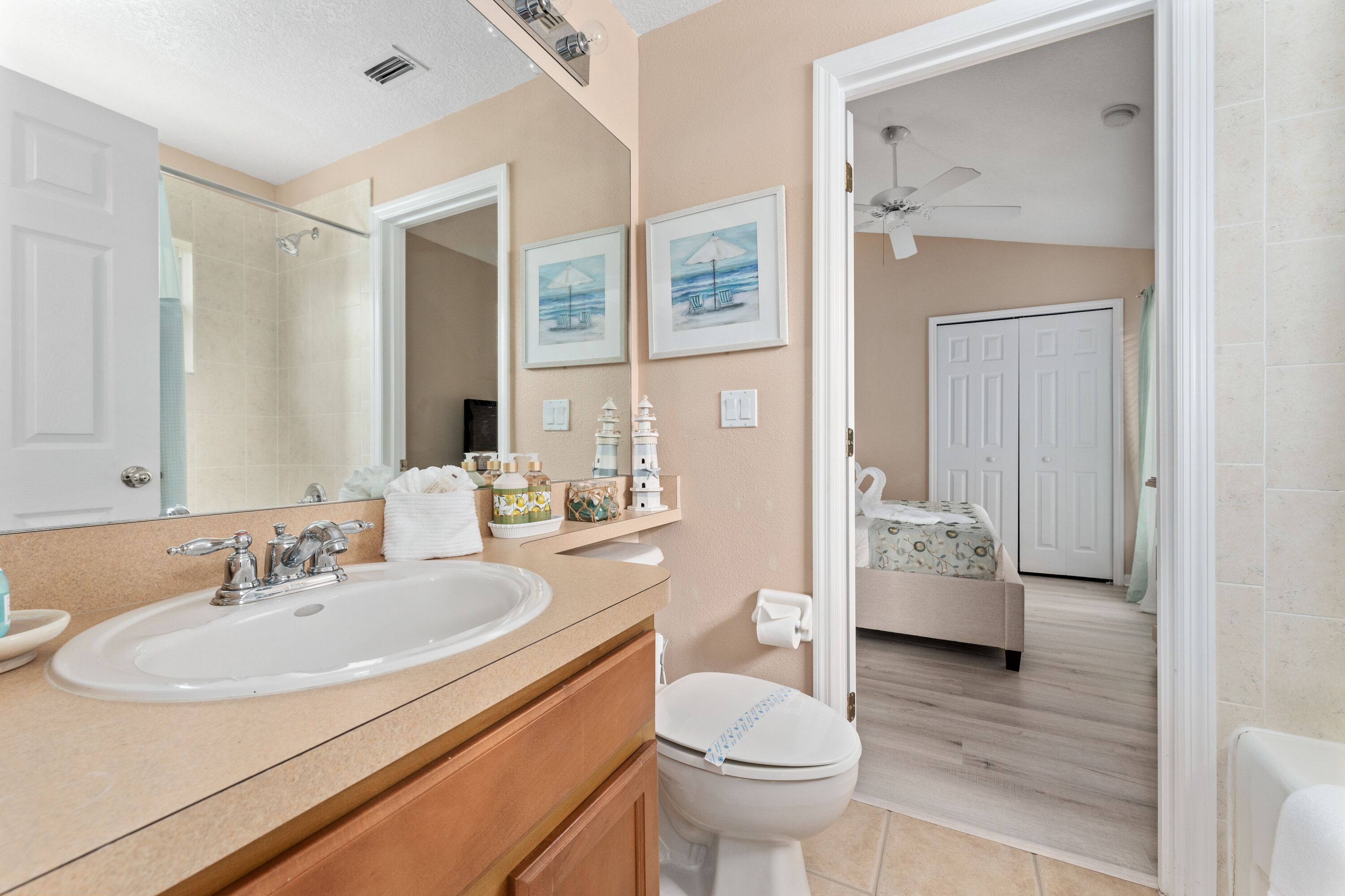A bathroom with a double sink, a toilet, and a shower. There are framed pictures on the wall and a ceiling fan.