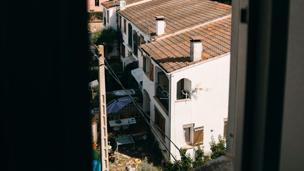 Blick durch ein Fenster auf ein Gebäude mit Ziegeldach und sichtbaren Balkonen, bewölkter Himmel.