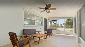 Een zonnedek met een plafondventilator, een bank, twee fauteuils, een kleine tafel en een groot raam met uitzicht op de omgeving.