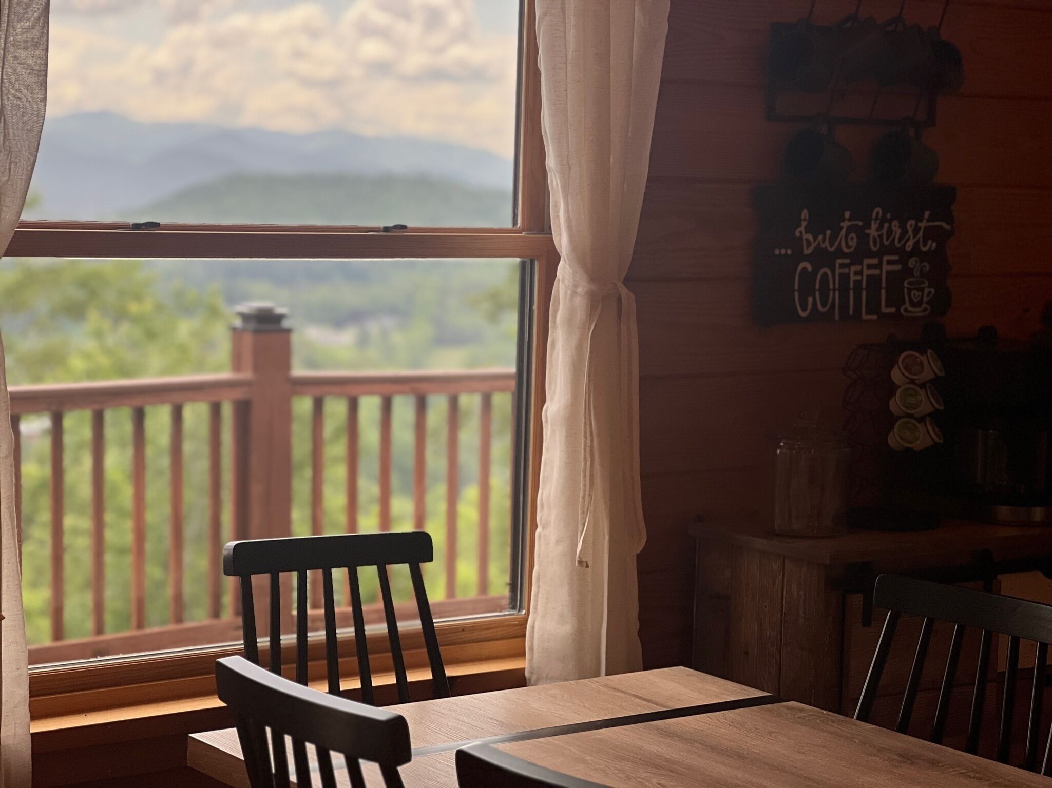 Une table et des chaises en bois près d’une fenêtre donnant sur une terrasse et des montagnes. Une pancarte sur le mur indique : « mais avant tout, du CAFÉ ».