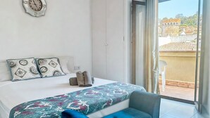 Une chambre à coucher avec un lit, des oreillers, un banc, une horloge et une vue sur un immeuble par la fenêtre.