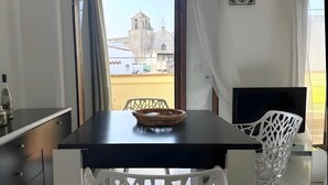 Un balcon donnant sur une église, une table à manger avec un panier, des chaises blanches et un téléviseur.