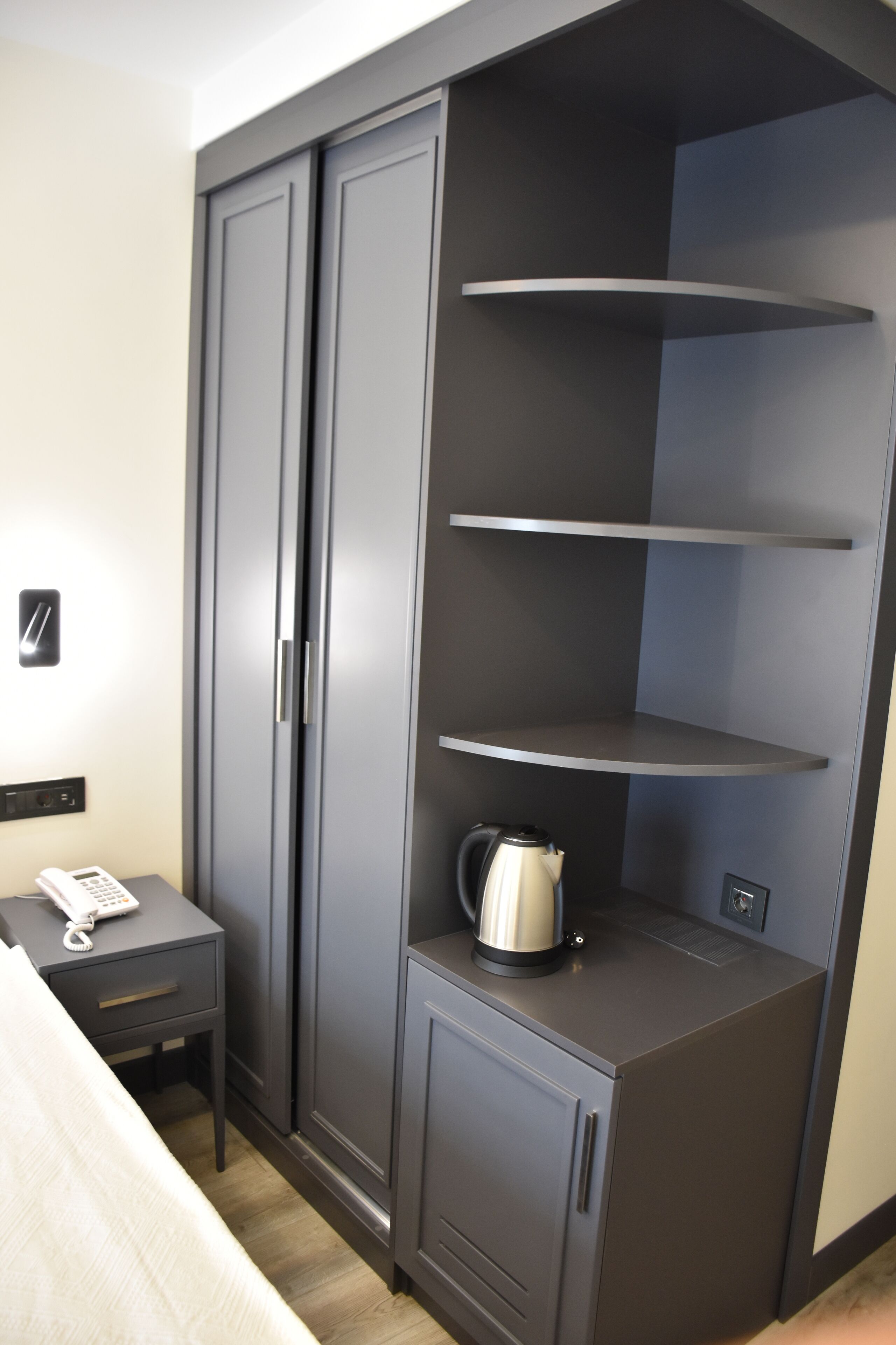 Una habitación con cama, mesita de noche con teléfono, armario con repisas y hervidor en la repisa inferior.