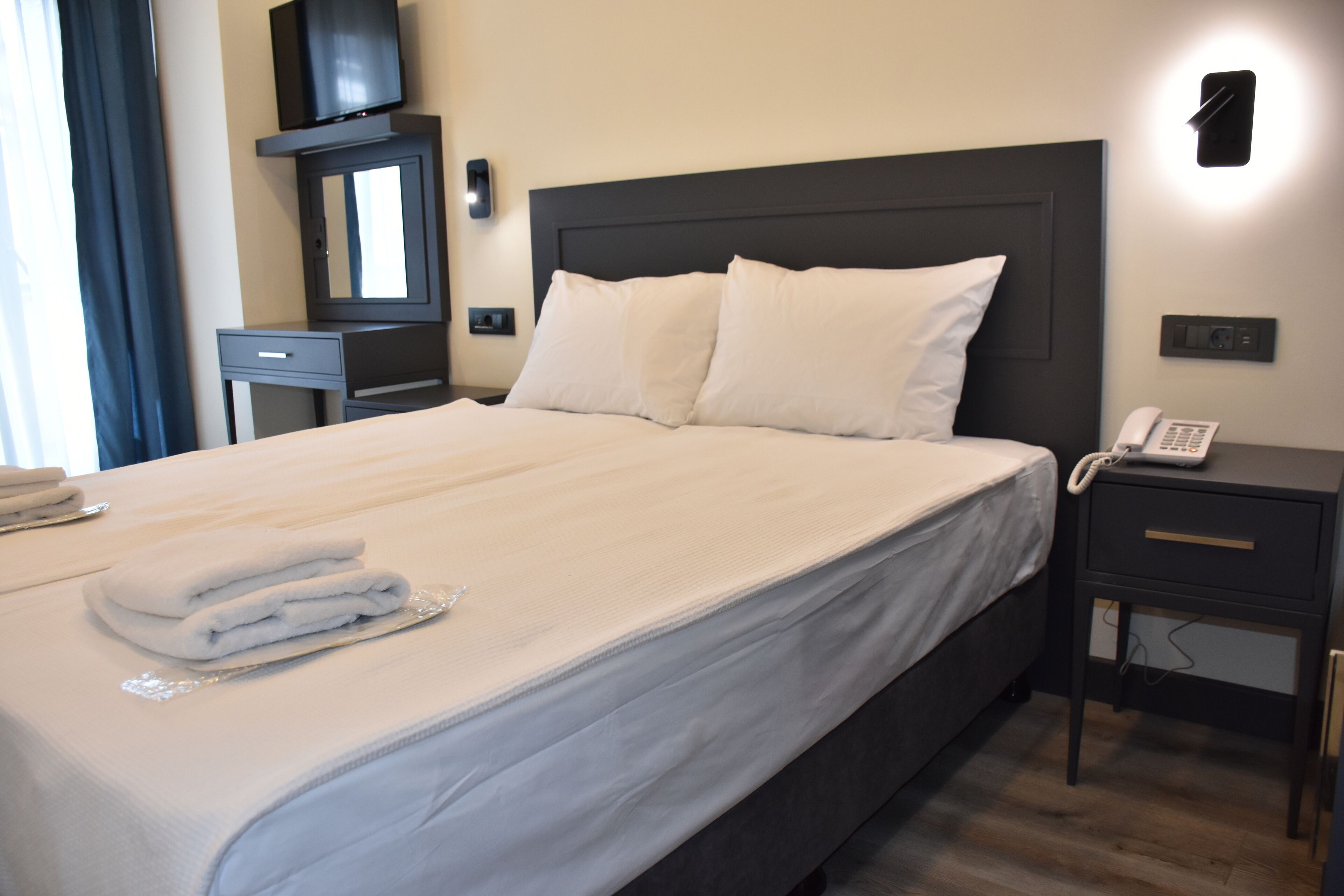 A neatly made bed with white linens, a bedside table with a phone, and a small desk with a mirror and television.