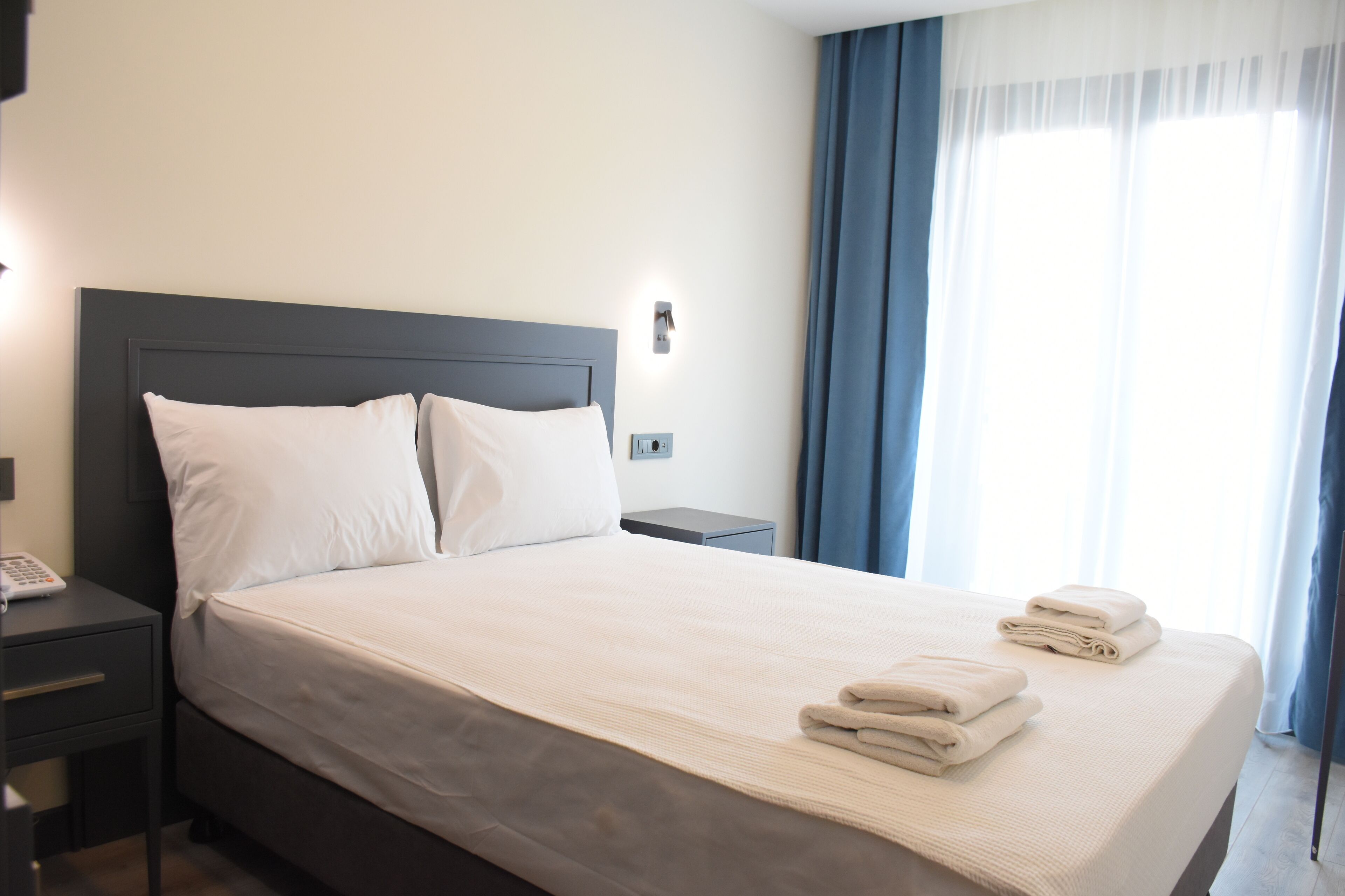 A neatly made bed with white linens and folded towels, a bedside table with a phone, and a nightstand with a lamp.