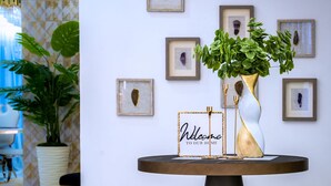 A round table with a 'Welcome to our home' sign, a gold vase with green plants, and framed artwork on the wall.