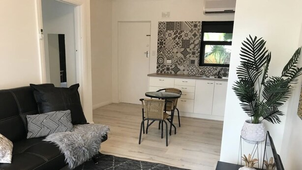 A modern living room with a black leather sofa, patterned rug, and a dining area with a small round table and chairs. - Wisteria Place (Plympton)