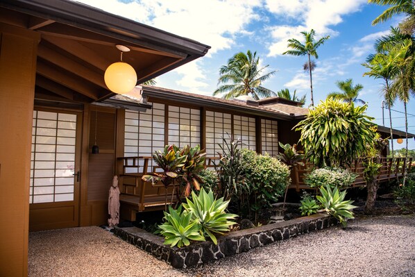 Ein traditionelles japanisches Gebäude mit Holzfassade, einem Kachelbelag am Eingang und einem Garten mit unterschiedlichen Pflanzen und einem Kiesweg.
