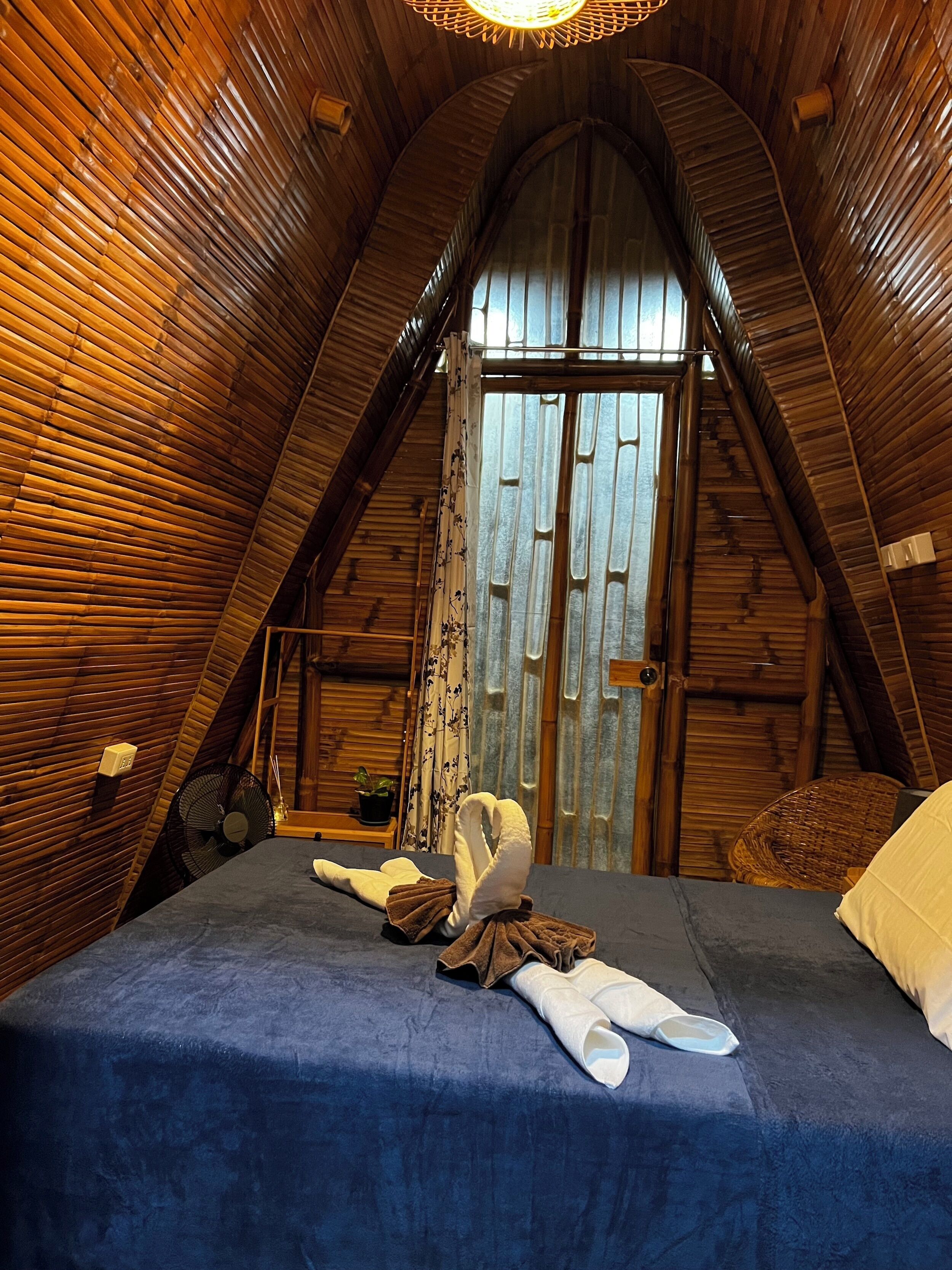 A cozy, wooden-paneled room with a bed, a fan, and a window with curtains.