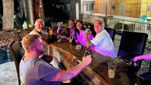 A group of people sitting around a wooden bar table with drinks, in an outdoor setting with a rustic wooden structure and a tiled wall in the background. - The Backyard Inn (Moalboal)