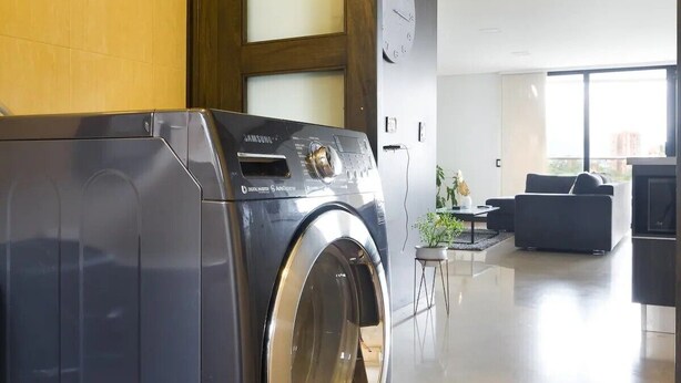 Un moderno cuarto de lavado con una lavadora Samsung, paneles de madera, un reloj grande, y una zona de estar minimalista con un sofá y una mesita de centro.