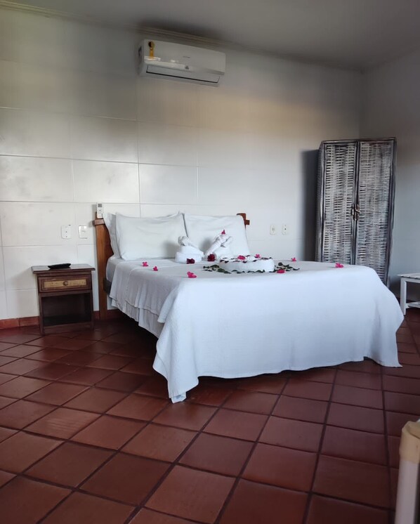 A neatly made bed with white linens and pink flowers, a small wooden nightstand with a bowl, and a wicker wardrobe. - Fantasias do Agreste (Jandaira)