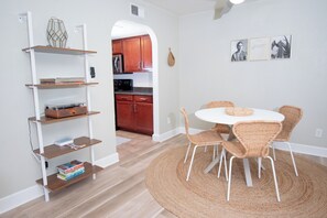 A dining area with a round table and wicker chairs, a bookshelf with books and decorative items, and a kitchen visible through an archway. - Endless CASAbilities at this updated, clean Arcadia condo (Phoenix)