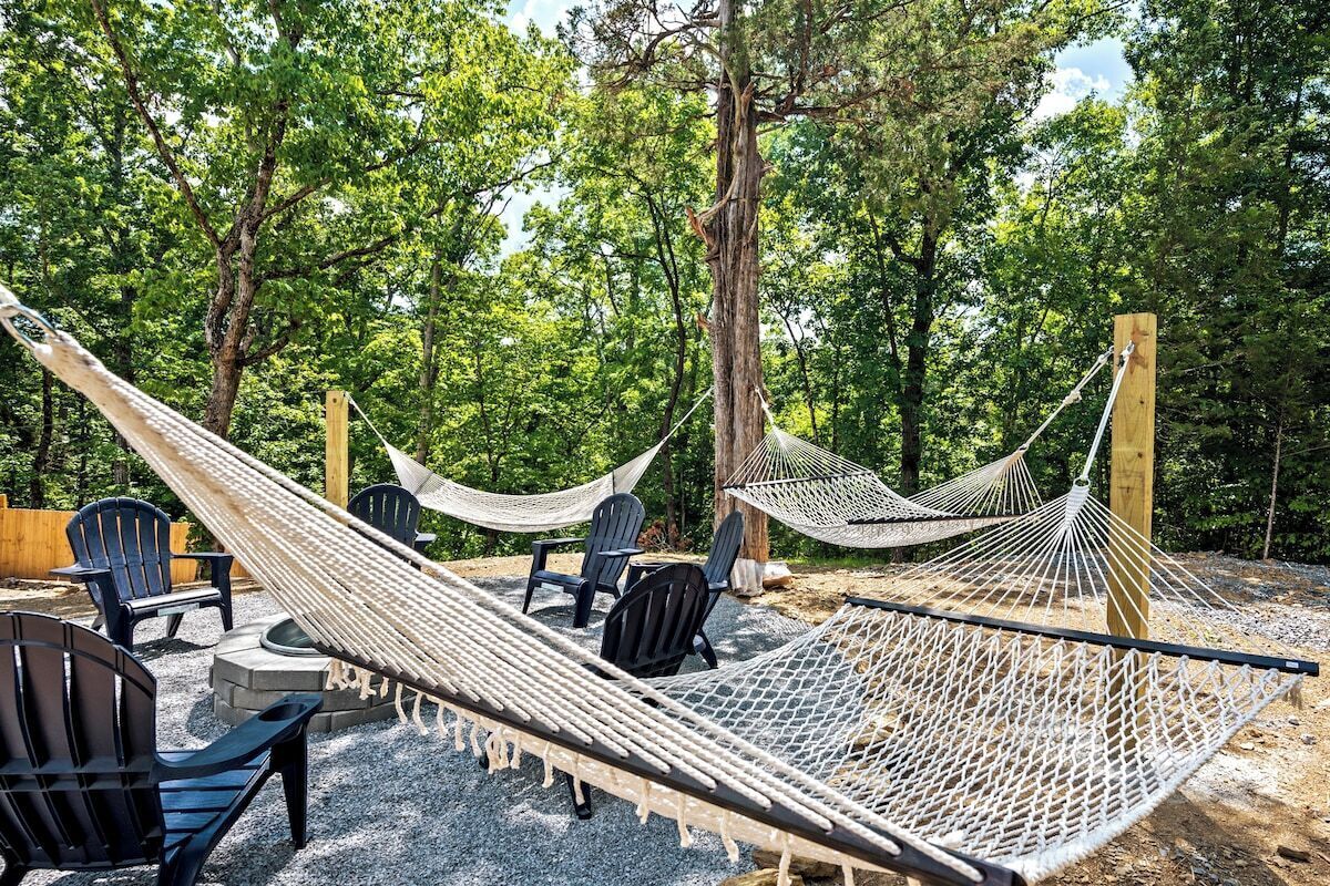 Un hamac est installé entre des arbres dans une zone boisée, avec des fauteuils Adirondack et un foyer.