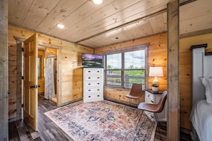 Interior de una cabaña de madera con una cama, un escritorio con lámpara, un televisor y una alfombra estampada.