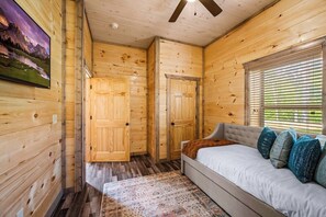 Een houten blokhut van binnen, met een bed, een plafondventilator en een raam met zonwering.