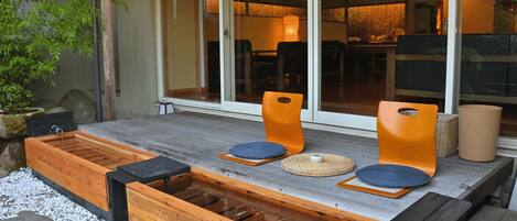 A wooden deck with two orange chairs and a small table, a wooden bench, and a water feature.