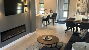 Una sala de estar moderna con un televisor de pantalla plana grande, una chimenea, un sofá oscuro y un comedor con mesa y sillas.