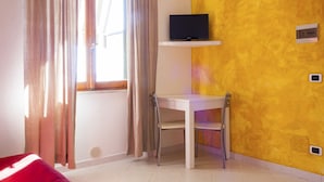 Une pièce comprenant un bureau, une chaise, un téléviseur posé sur une étagère, un canapé rouge et une fenêtre avec des rideaux.