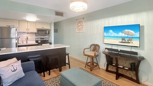 Une cuisine moderne équipée d’un réfrigérateur, d’un four à micro-ondes et d’une cuisinière, un coin repas avec un bar, un salon avec un canapé et un pouf, et un grand téléviseur fixé au mur.