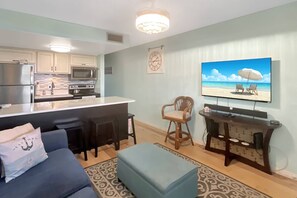 Une cuisine moderne équipée d’un réfrigérateur, d’un four à micro-ondes et d’une cuisinière, un coin repas avec un bar, un salon avec un canapé et un pouf, et un grand téléviseur fixé au mur.