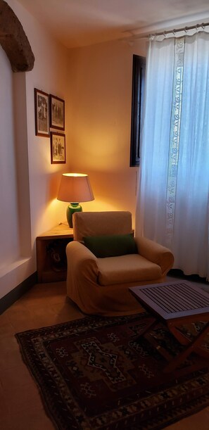 Un rincón acogedor con un sillón beige, un cojín verde, un tapete estampado, una lámpara de mesa y una ventana con cortinas de encaje.