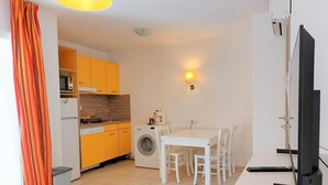 A compact kitchen with a microwave, sink, and washing machine, a dining area with a table and chairs, and a TV on a stand. - Petite Maison (Belgodère)