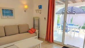 A living room with a sofa, a coffee table, a framed picture, a wall-mounted lamp, and a sliding glass door leading to an outdoor patio area with a table and chairs. - Petite Maison (Belgodère)
