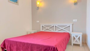 A bedroom with a red bedspread, white headboard, two bedside tables, and wall-mounted lights. - Petite Maison (Belgodère)