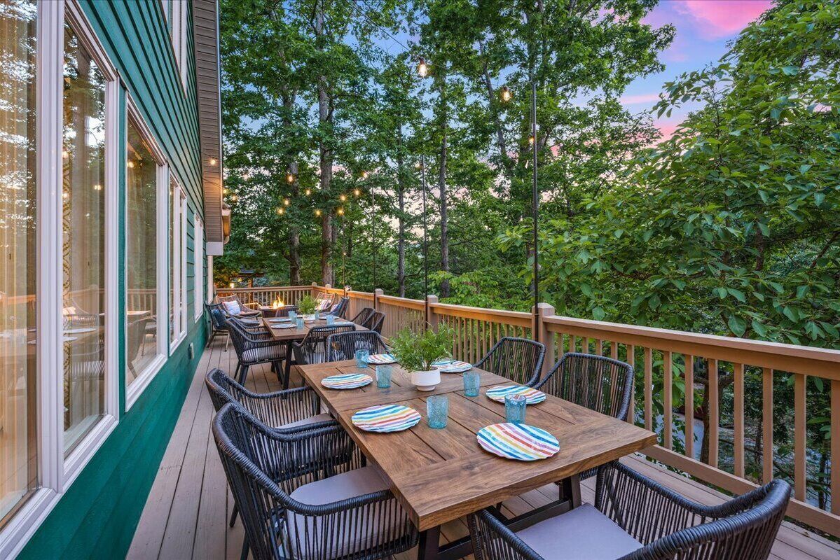 An outdoor dining area with wooden tables and chairs, string lights, and a view of trees and sky.