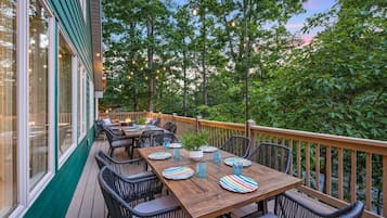 An outdoor dining area with wooden tables and chairs, string lights, and a view of trees and sky.