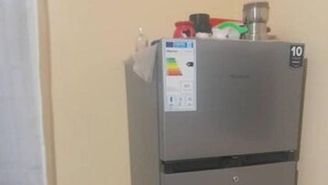 A compact, metallic refrigerator with a water dispenser, located in a small room with a single bed and a wall-mounted shelf. - Habesha circle (kampala)