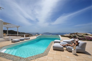 Ein Poolbereich mit gewölbter Kante, Sitzmöglichkeiten im Freien und Blick auf das Meer.