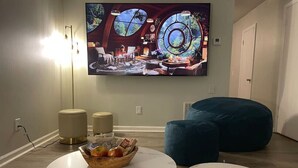 Una sala de estar con un televisor de pantalla plana montado en la pared, dos mesas redondas, un sillón de peluche y un frutero sobre una de las mesas.