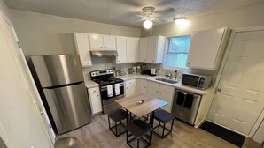 Cocina compacta con gabinetes blancos, refrigerador de acero inoxidable, estufa a gas, microondas y una pequeña mesa de comedor con cuatro sillas.