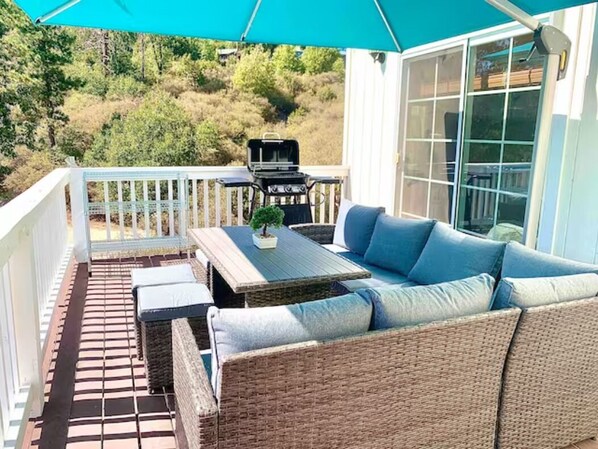 Relax on the patio with a wicker sofa, small table, and grill, surrounded by a wooden deck and a scenic view.