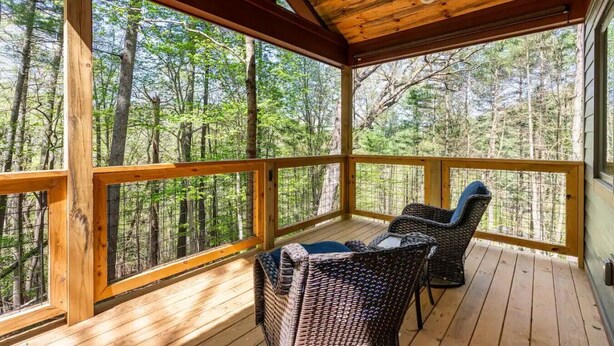 Um deck de madeira com cadeiras de vime e um ventilador de teto, com vista para uma área arborizada.