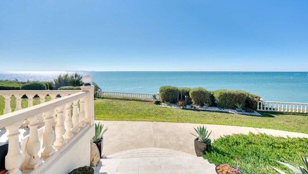 Un balcón con vista al mar, barandilla blanca y zona pavimentada con plantas en macetas.