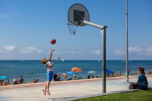Un panier de basketball installé sur une plage, offrant une vue dégagée sur l’océan et où l’on aperçoit des gens se détendant à l’arrière-plan.
