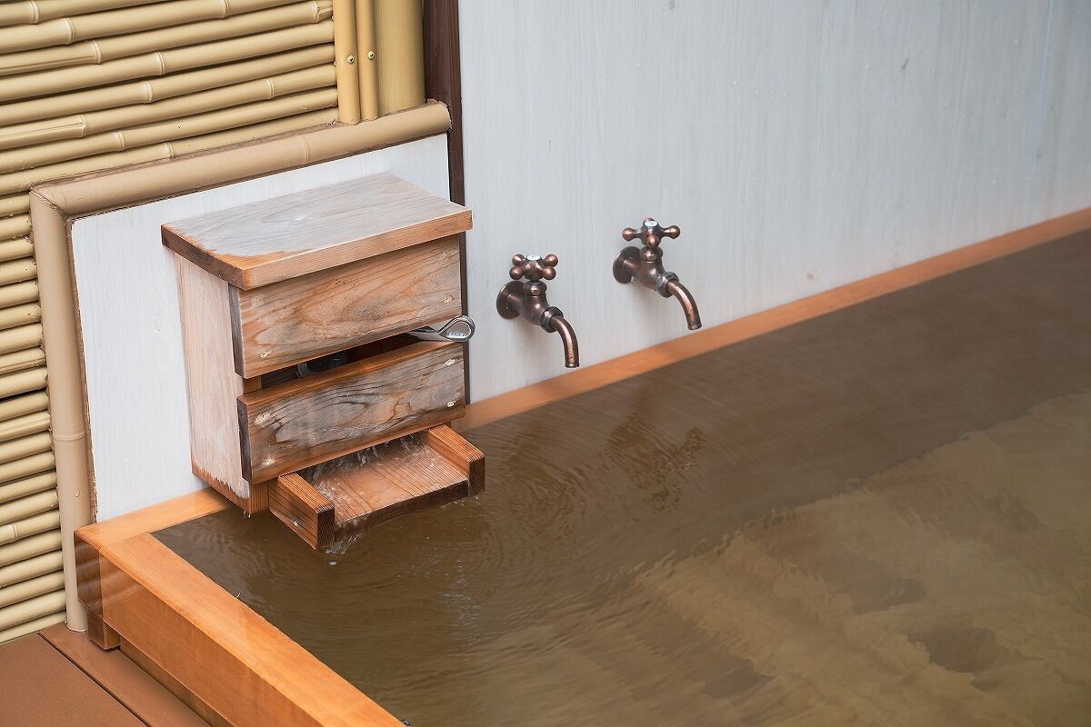 A wooden bench with a built-in shelf and a small wooden table next to a wall with two faucets.