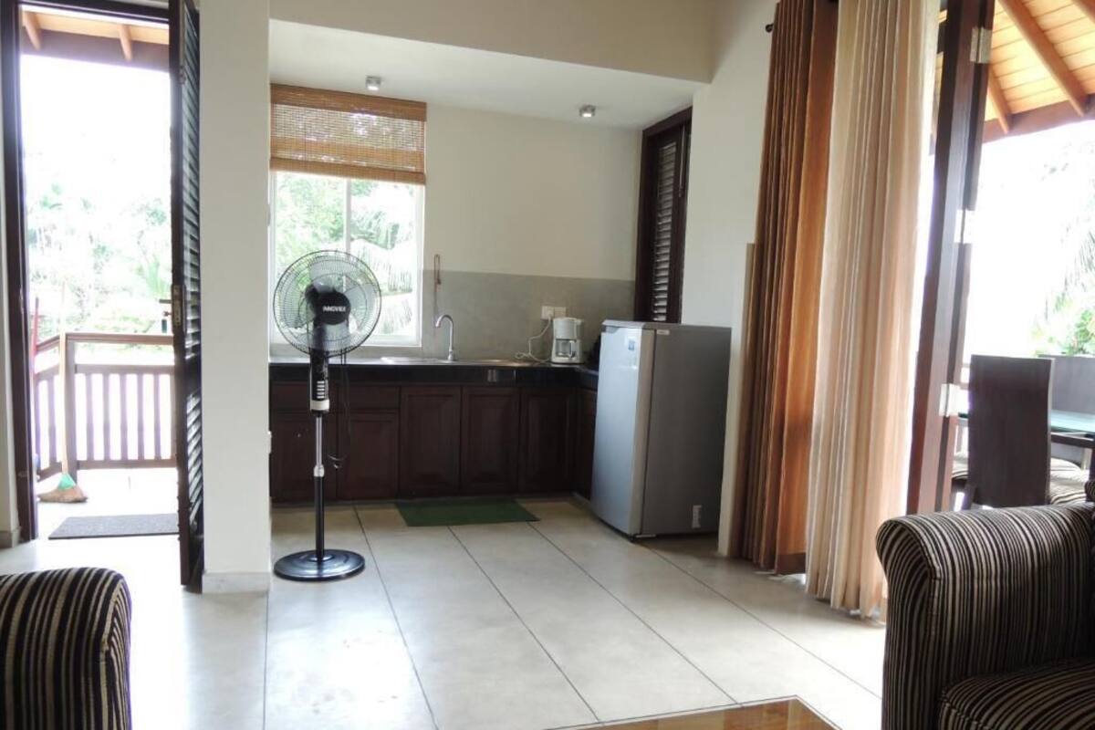 A living room with a sofa, a coffee table, a fan, and a kitchen area with a refrigerator and a coffee maker.