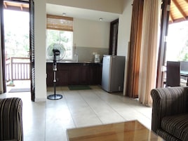 Una sala de estar con un sofá, una mesa de centro, un ventilador y un área de cocina con un refrigerador y una cafetera.