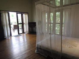 Una habitación con suelo de madera, una cama con dosel, un televisor y un ventanal corredero que da a un espacio exterior.